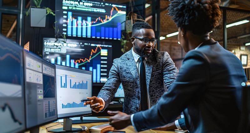 Two business professionals in formal attire discussing financial data on computer screens in a modern office environment. The background features multiple large monitors displaying stock market charts, bar graphs, and line graphs. One individual is pointing to data on a screen, while the other listens attentively. The setting reflects a high-tech financial or investor relations workspace.